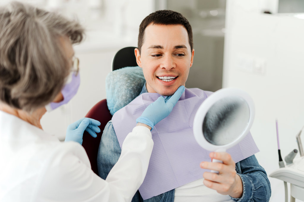 Smiling attractive male patient holding mirror, looking, talking to senior professional dentist. Concept of dental care, treatment, whitening